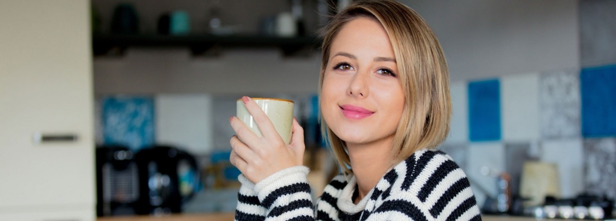 Jonge dame in trui zit aan tafel tijdens het ontbijt met een kop koffie in de keuken.